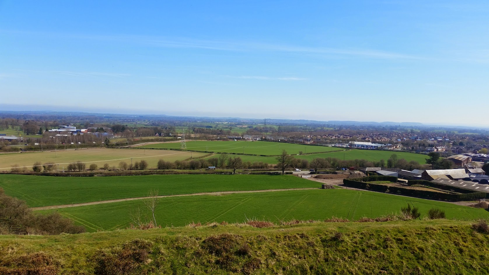 A picture from the top of a hill (a local hillfort). Verdant green fields stretch off into the distance. The sky is blue, with some wispy white clouds; we can see farm buildings to our right, and there is a small town just in view in the distance. There are hills just on the horizon.
